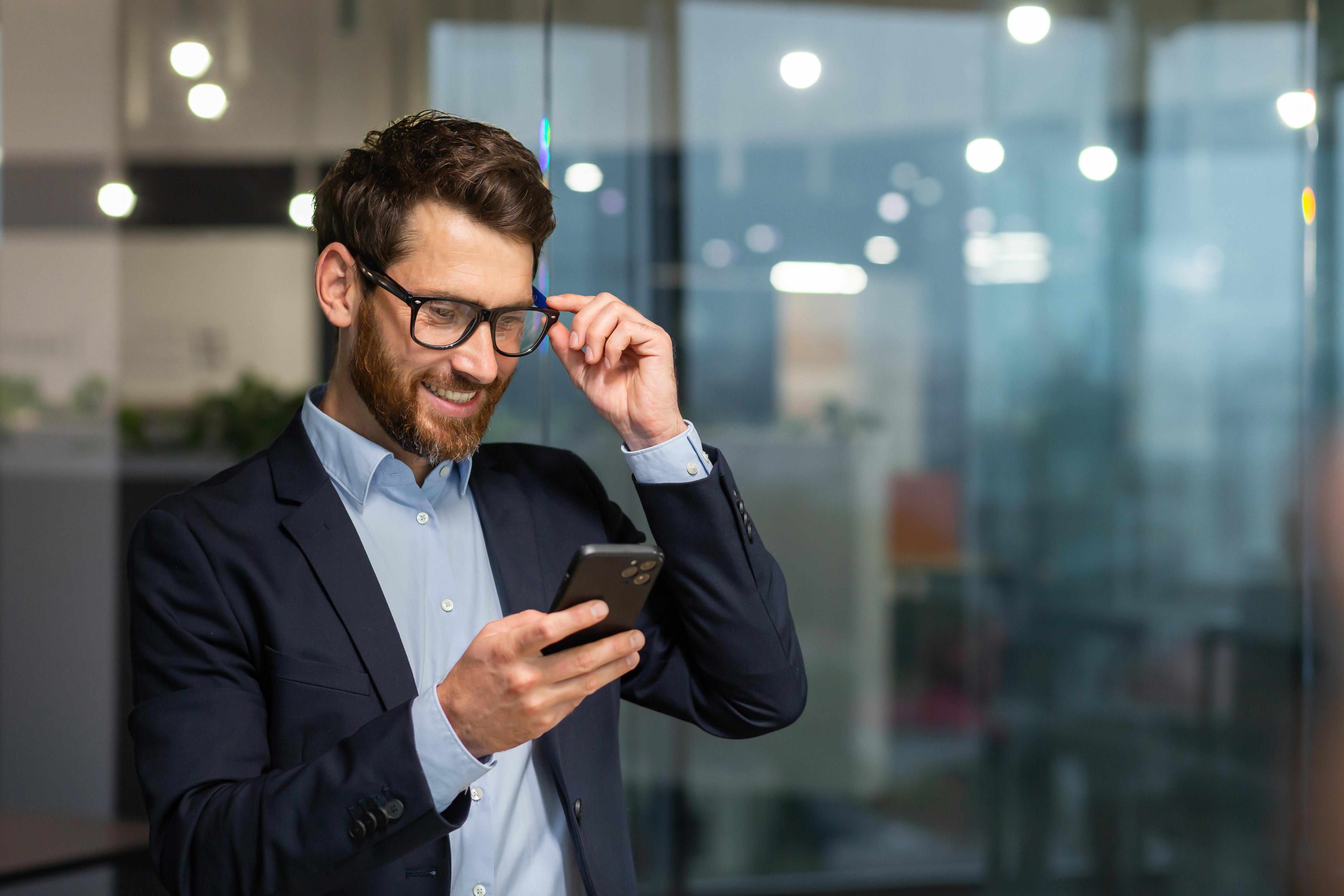 Successful financier investor works inside office at work, businessman in business suit uses telephone near window, man smiles and reads good news online from smartphone.